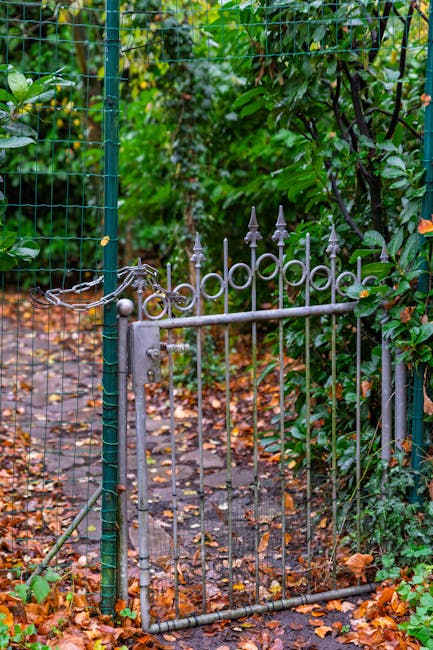 A small, decorative wrought iron garden gate with pointed finials at the top, painted in a weathered silver-grey color, partially open within a lush garden setting. The gate is connected to a green me