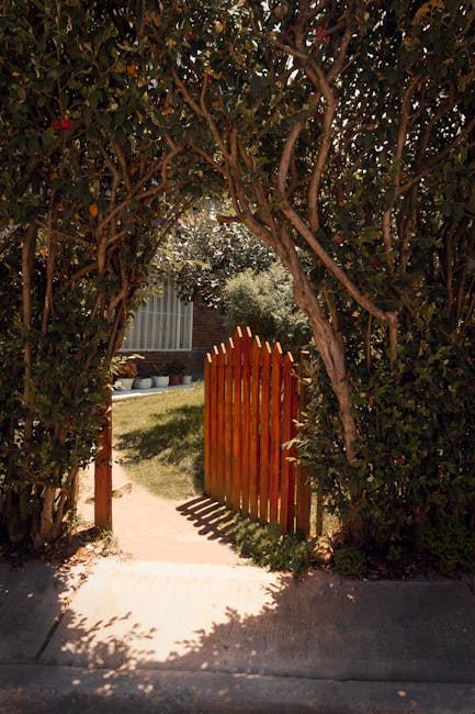 A small, decorative wrought iron garden gate with pointed finials at the top, painted in a weathered silver-grey color, partially open within a lush garden setting. The gate is connected to a green me